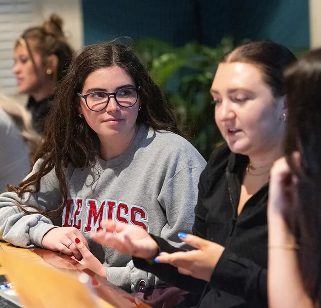 Three students gathered, seated; one looking intently while other speaks. Person in focus wears a sweatshirt that prominently says "Ole Miss"