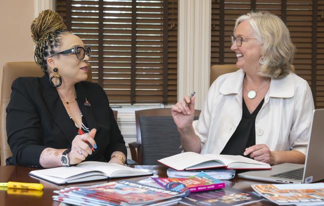 Kirsten Dellinger and Val Ross seated at a table talking together. There is a pile of magazines and books on the table in front of them.