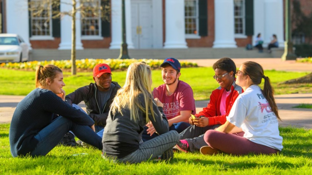 A small group of students are sitting in a circle on the grass in front of the Lyceum building at UM campus