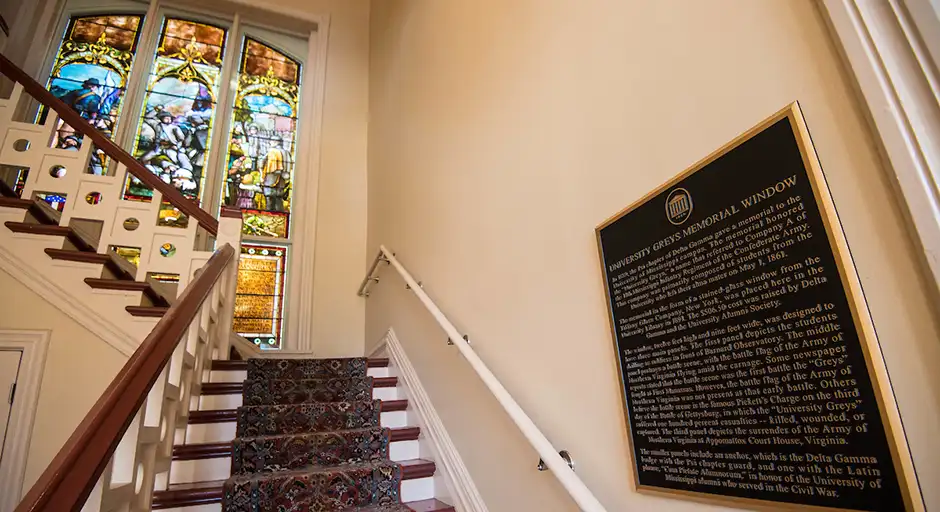 Staircase with colorful stained glass window on landing. Plaque on wall with paragraph of text that is not legible in the photo.