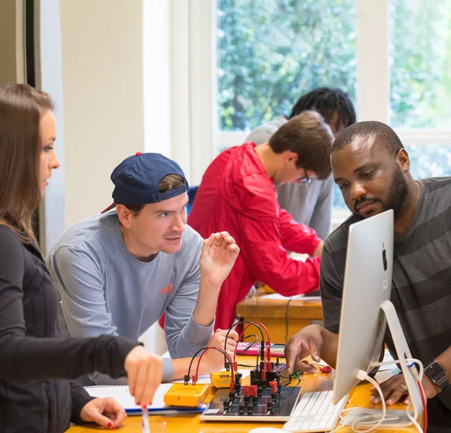 Two students and instructor in physics lab classroom gathered around a desktop computer and instructional model with wires