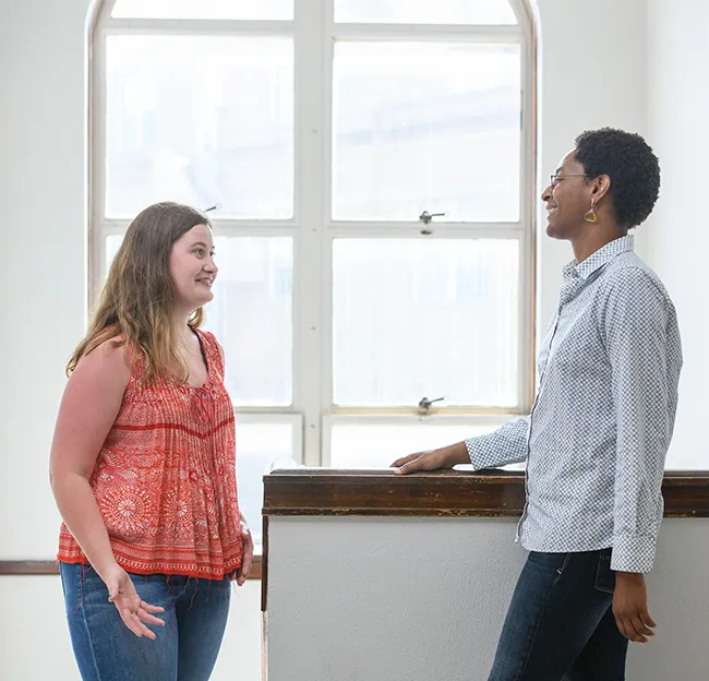 Student and Professor engaged in conversation in a white stairwell. Two people are in profile.