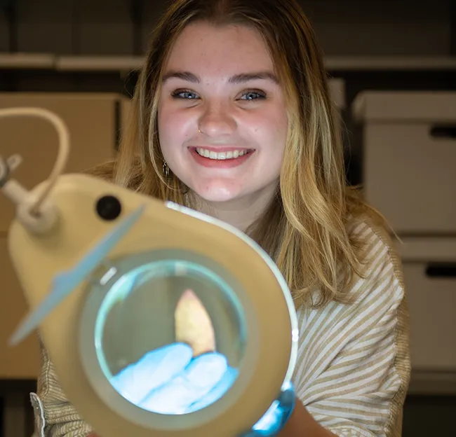 Person smiling, looking at camera holding an archaeological artifact that is under a magnifier in foreground