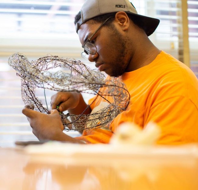 Man sitting at a table using a tool to bend a wire frame to make a sculpture of a turtle