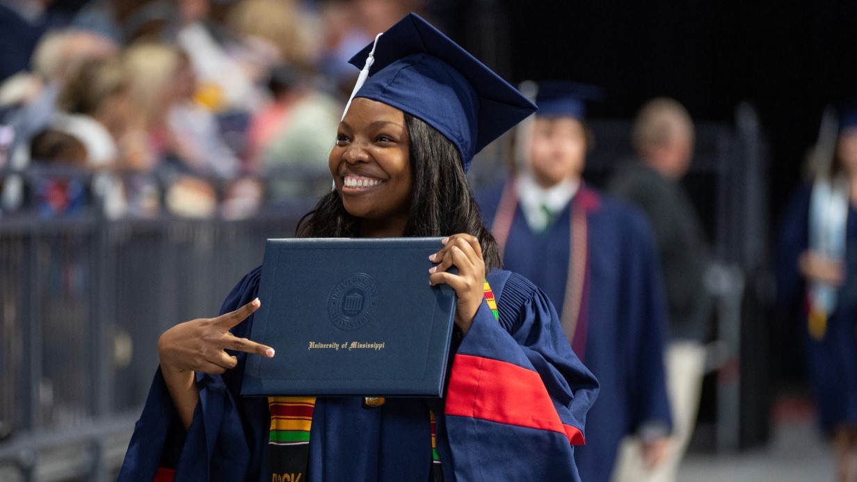 student holding diploma cover at graduation smiling at family in the stands and showing the "victory" sign