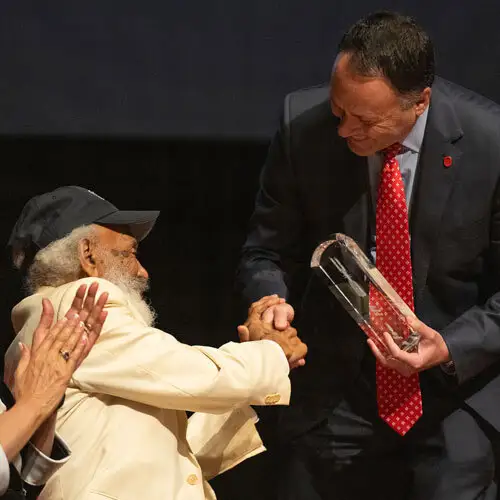 two men shaking hands. Person on left seated, wearing a baseball cap and white suit; person on right standing, bent over holding a crystal award