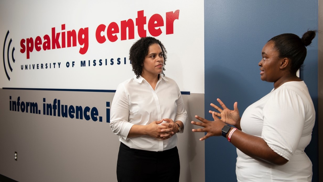 faculty member and student talking together next to a wall with a sign for the Speaking Center