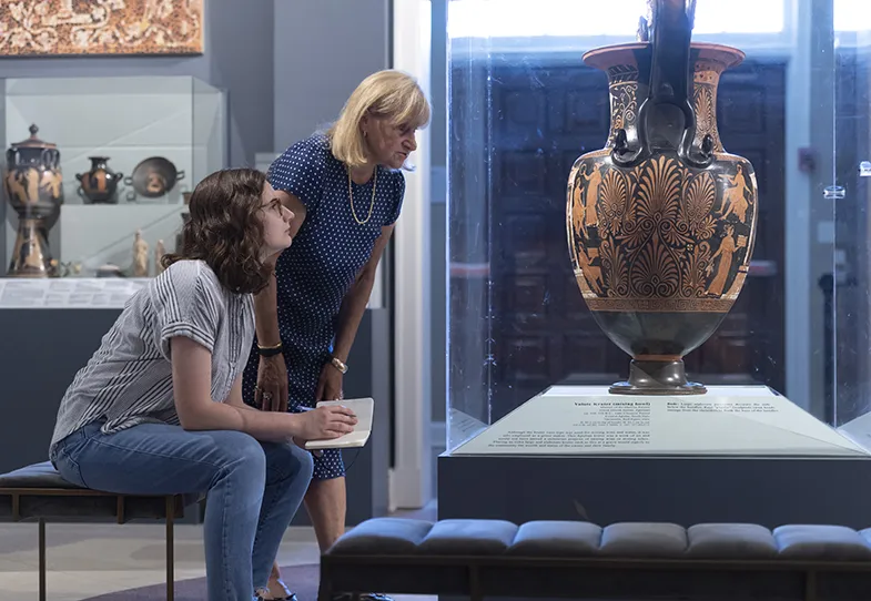 Professor (standing) and student (sitting) in museum setting looking at large Greek urn.