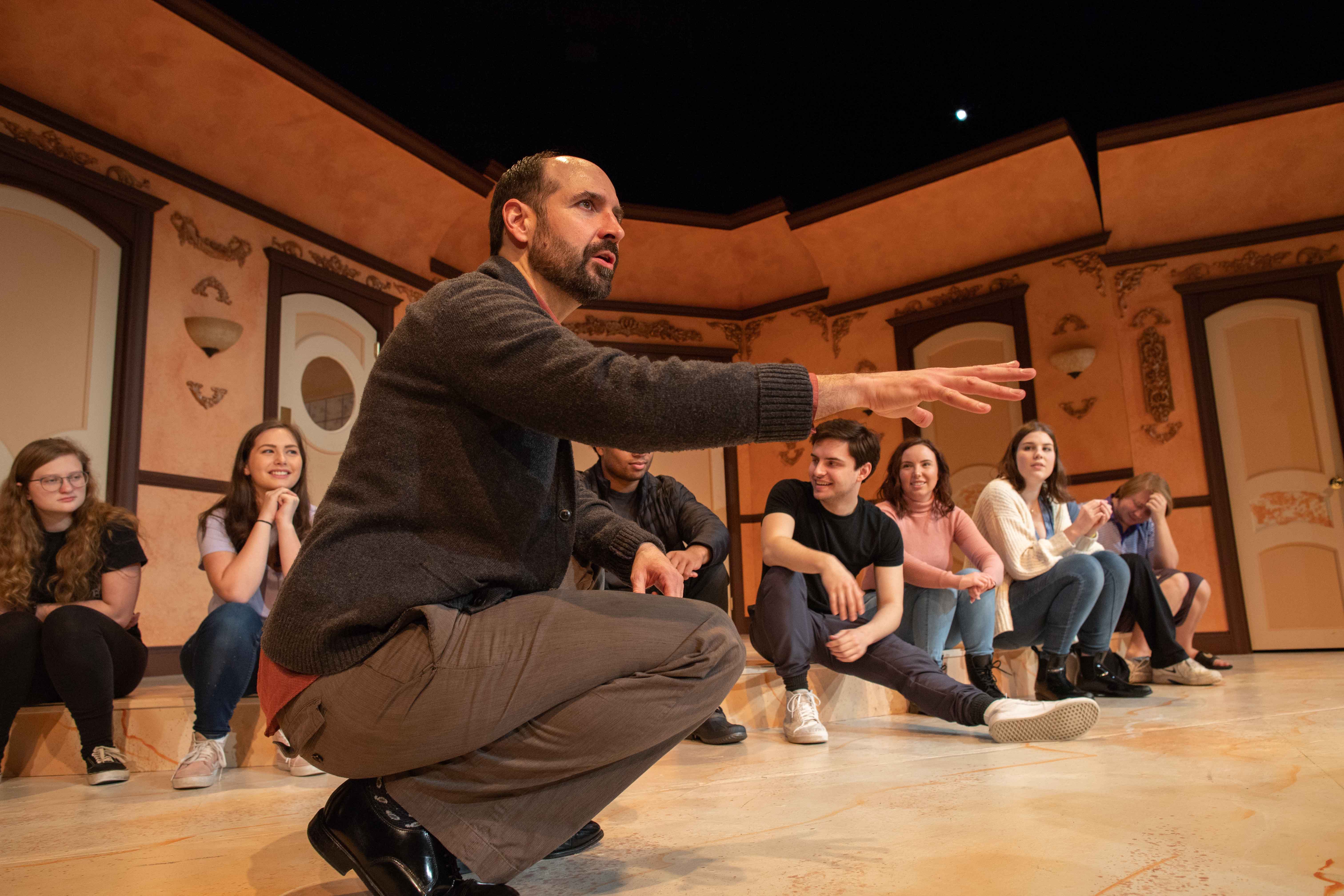 A professor kneels in front of students. He directs practice for a theatre production. Students sit behind him on stage. Behind the students is a stage set for an elegant house.