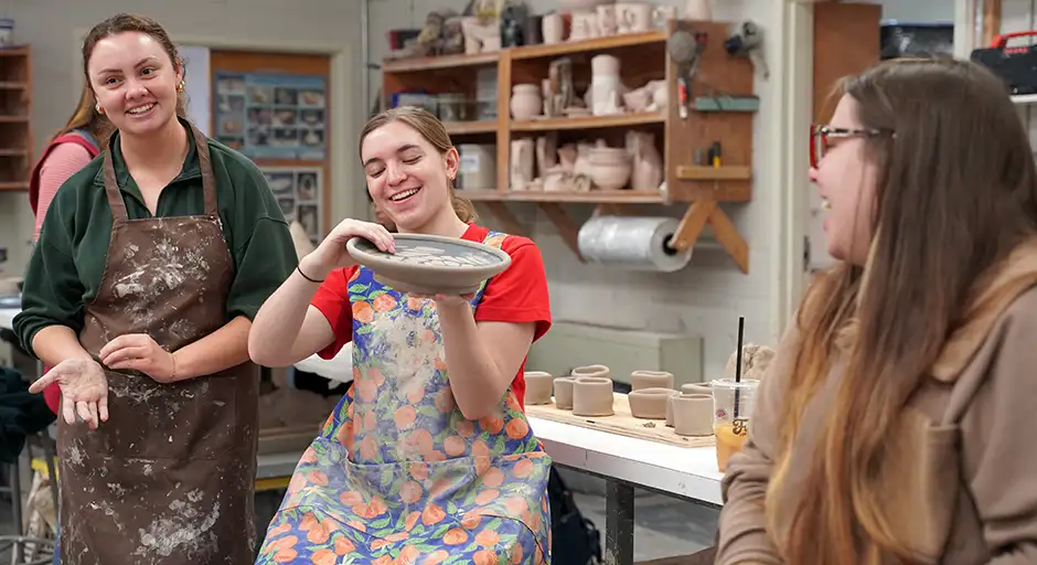 Three female students in the ceramics studio. They are smiling and laughing. One holds up a ceramic plate.