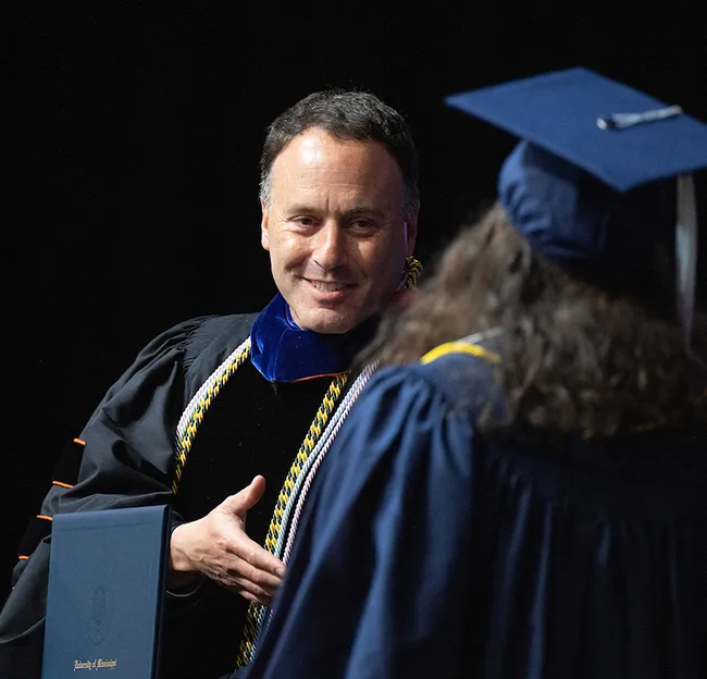 Dean Cohen in graduation regalia with hand out to shake graduate's hand.