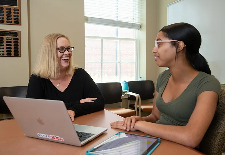 Smiling professor and student sitting at table engaged in conversation. Lap top computer opened and in foreground.