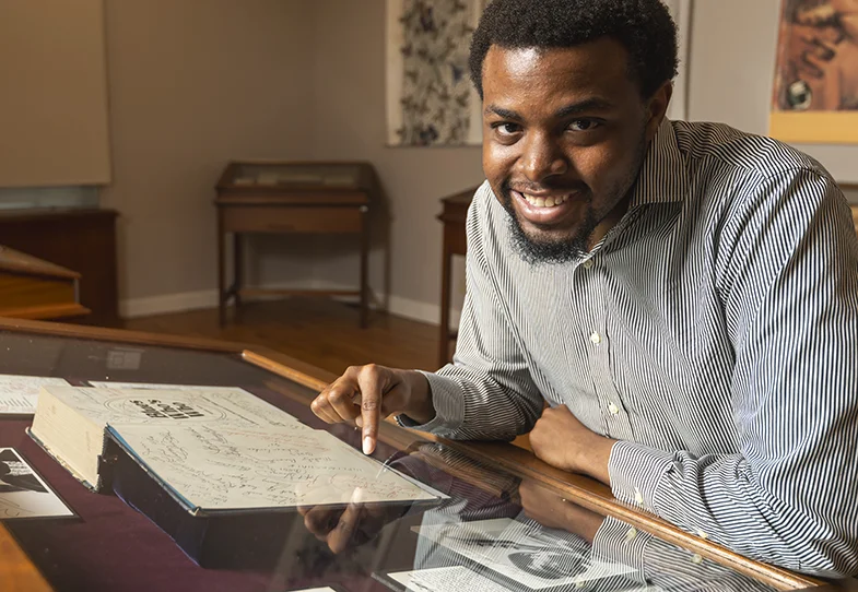 person sitting at a table; looking at the camera and pointing to a glass case holding archival books and letters