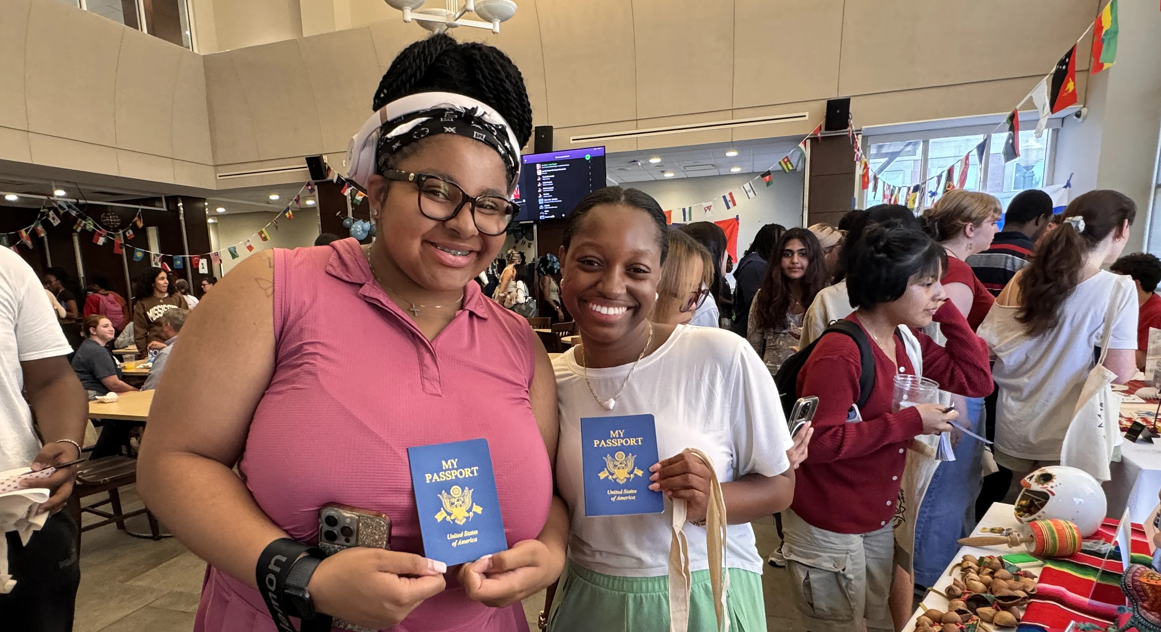 Two students hold up passports and smile at the camera. Lots of people milling around during a reception.
