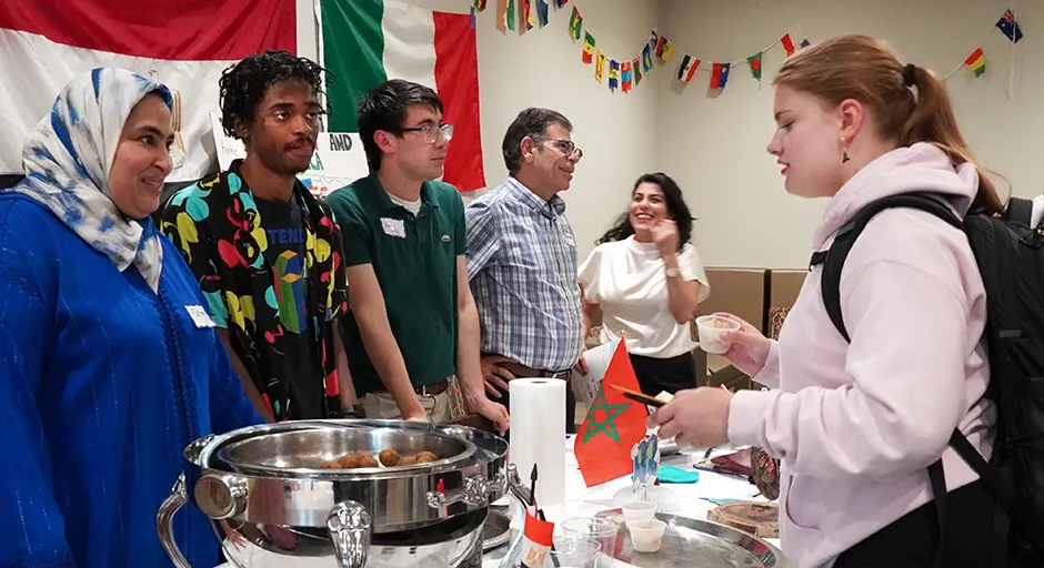 Students and professors stand around a table; flags hung in the background; metal serving dish with food is on the table
