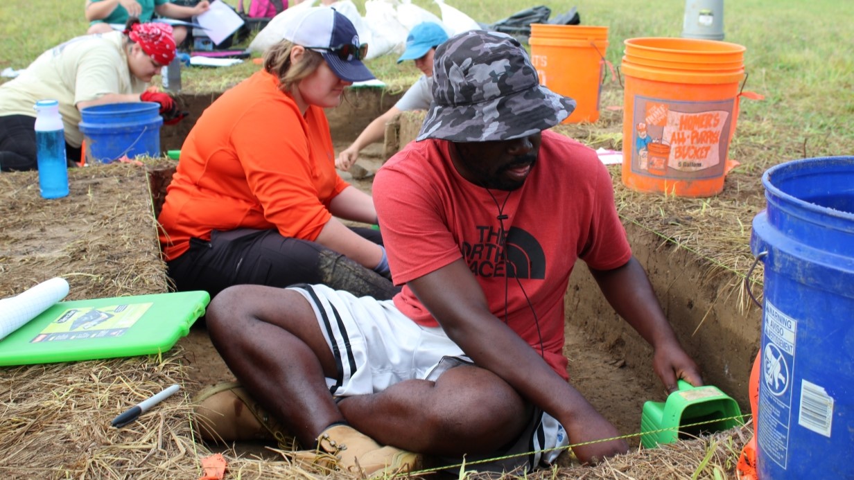 image of students sitting on the ground at an archaeology dig and they are digging in the ground