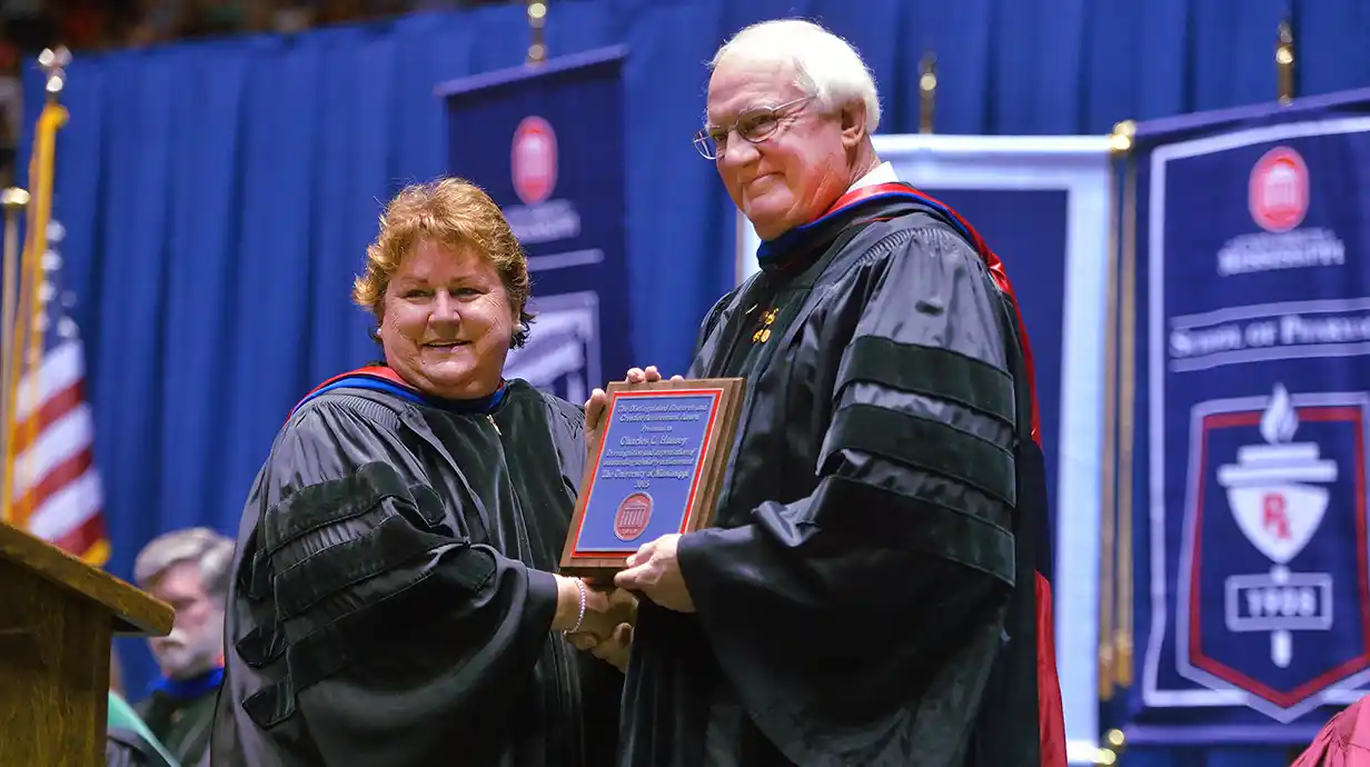 Two people on stage; one person is handing the other person a plaque. Both people are wearing regalia robes