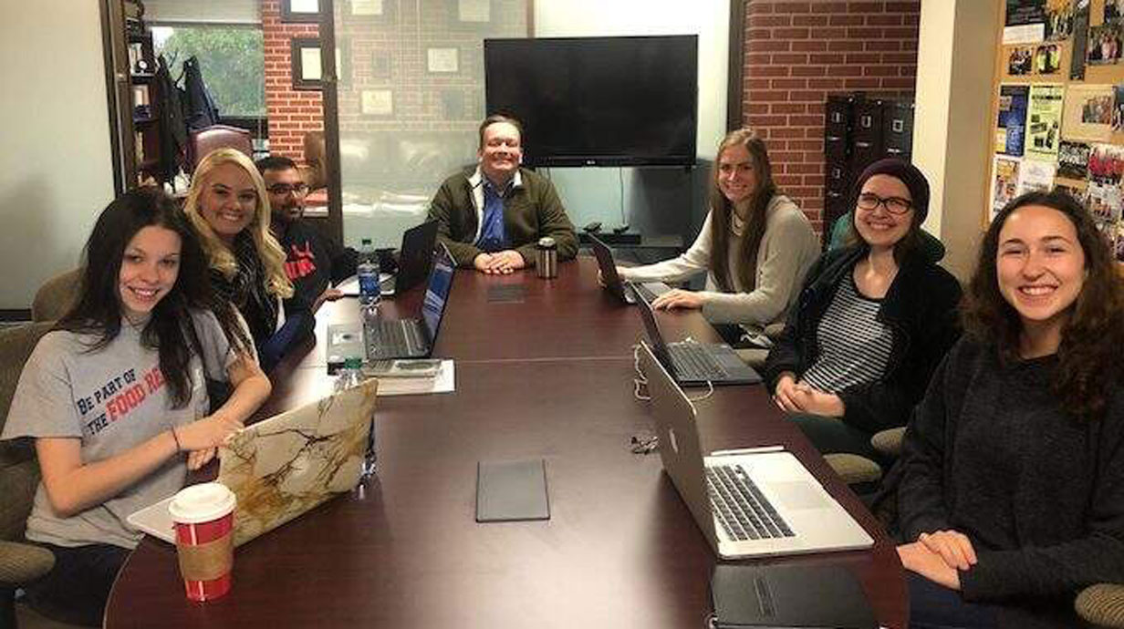 students and professor sit around a table and smile at the camera