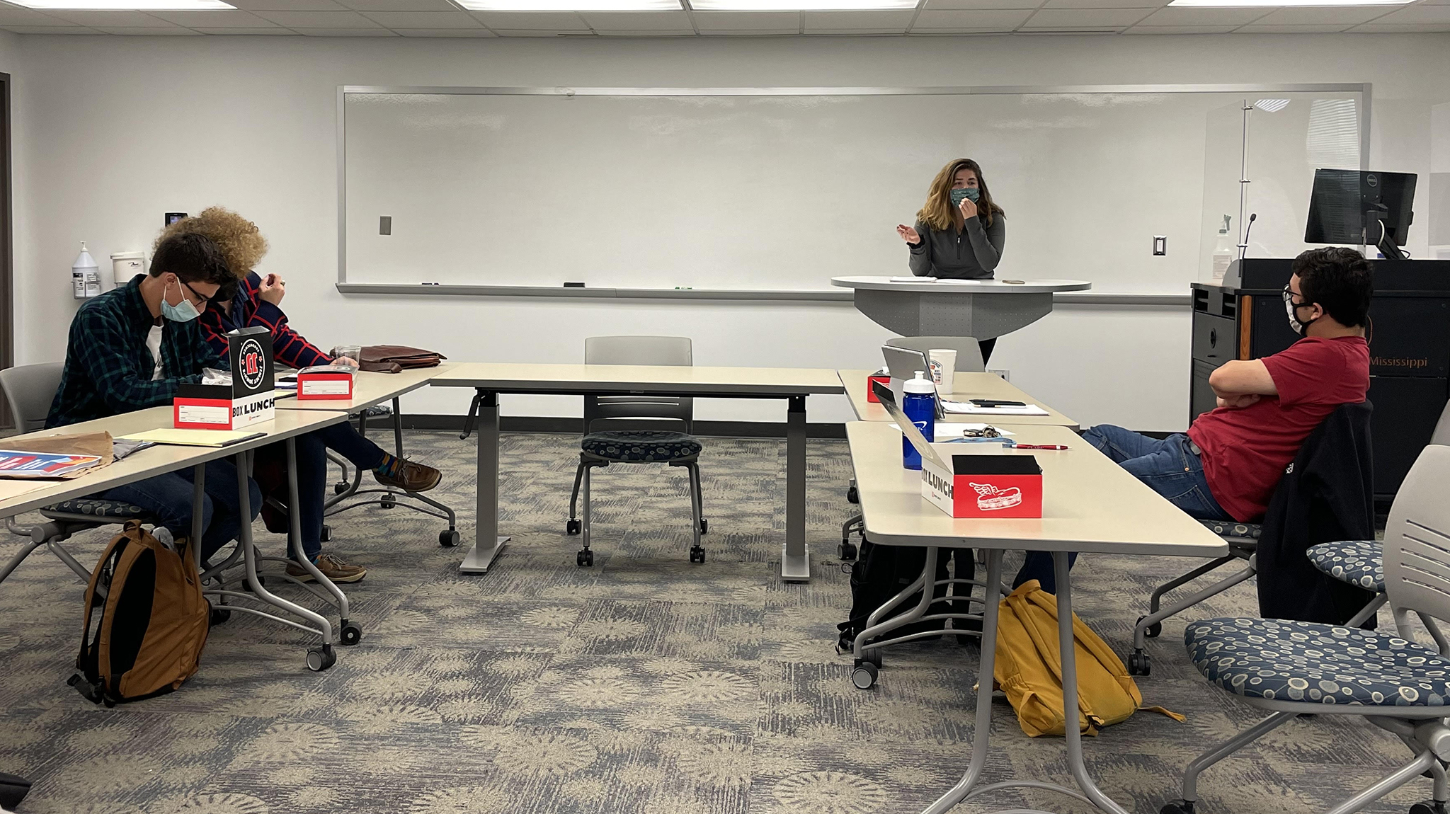 Four students in small classroom. Three are sitting at table looking at the fourth student standing and talking behind a podium.