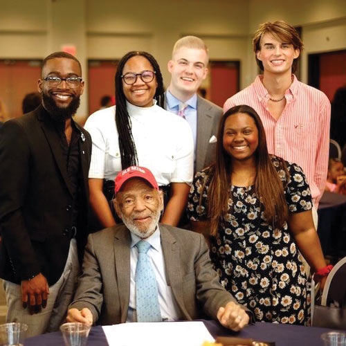 James Meredith seated at a desk wearing a red hat with students standing behind and around him.