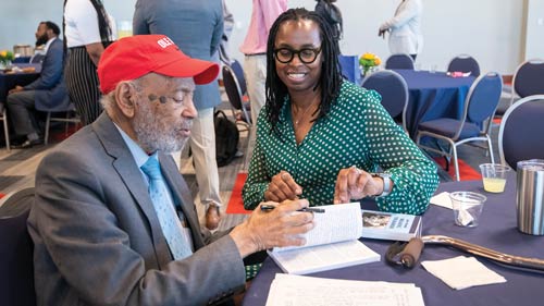 James Meredith sitting at a table signing his book with another person on his left.